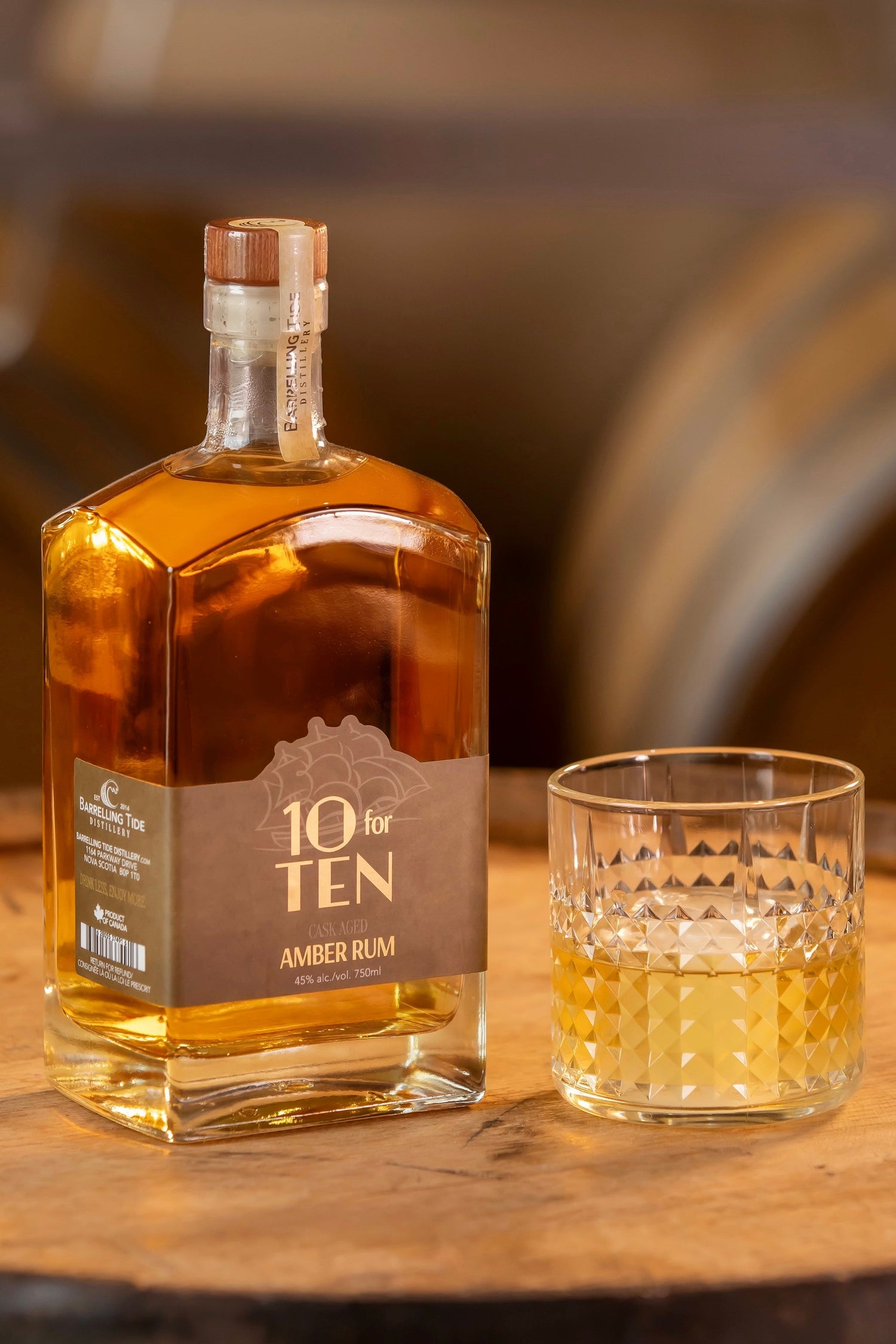 Bottle and glass of 10 for Ten Amber Rum on a wooden barrel with barrels in the background
