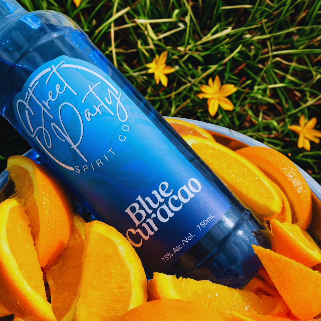 Bottle of Street Party Spirit Co. Blue Curacao surrounded by orange slices in a metal bucket on a grassy background
