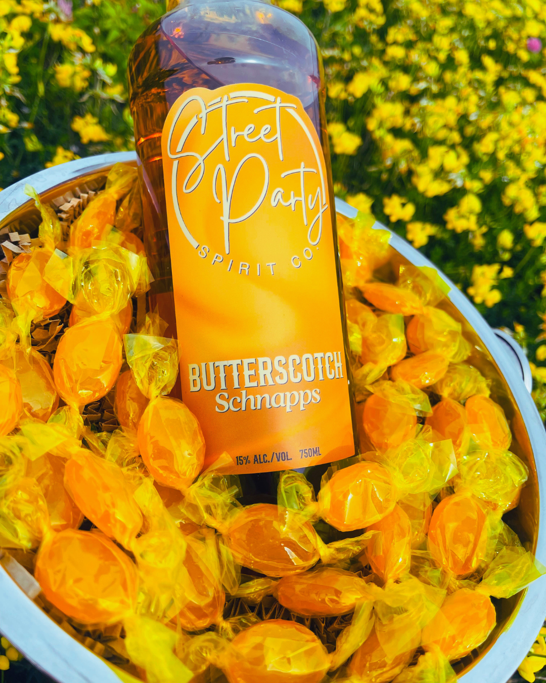 Bottle of Street Party Spirit Co. Butterscotch Schnapps sitting in butterscotch candy in a metal bucket against a floral background