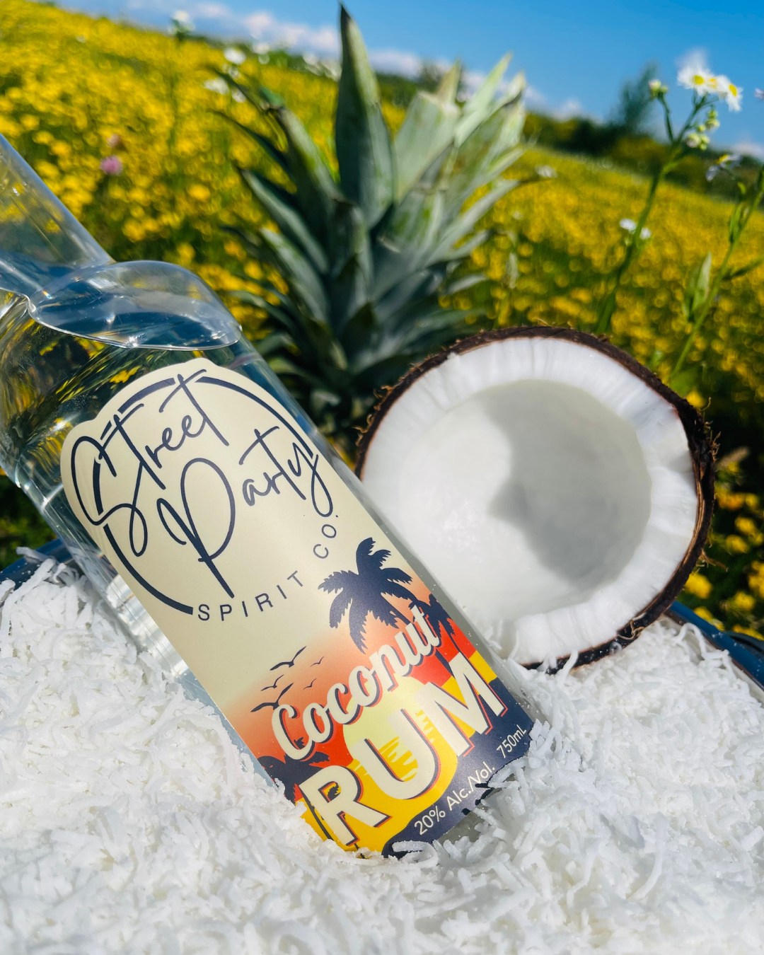 Bottle of Street Party Spirit Co. Coconut Rum with a pineapple and half a coconut on shreds of coconut in a field.