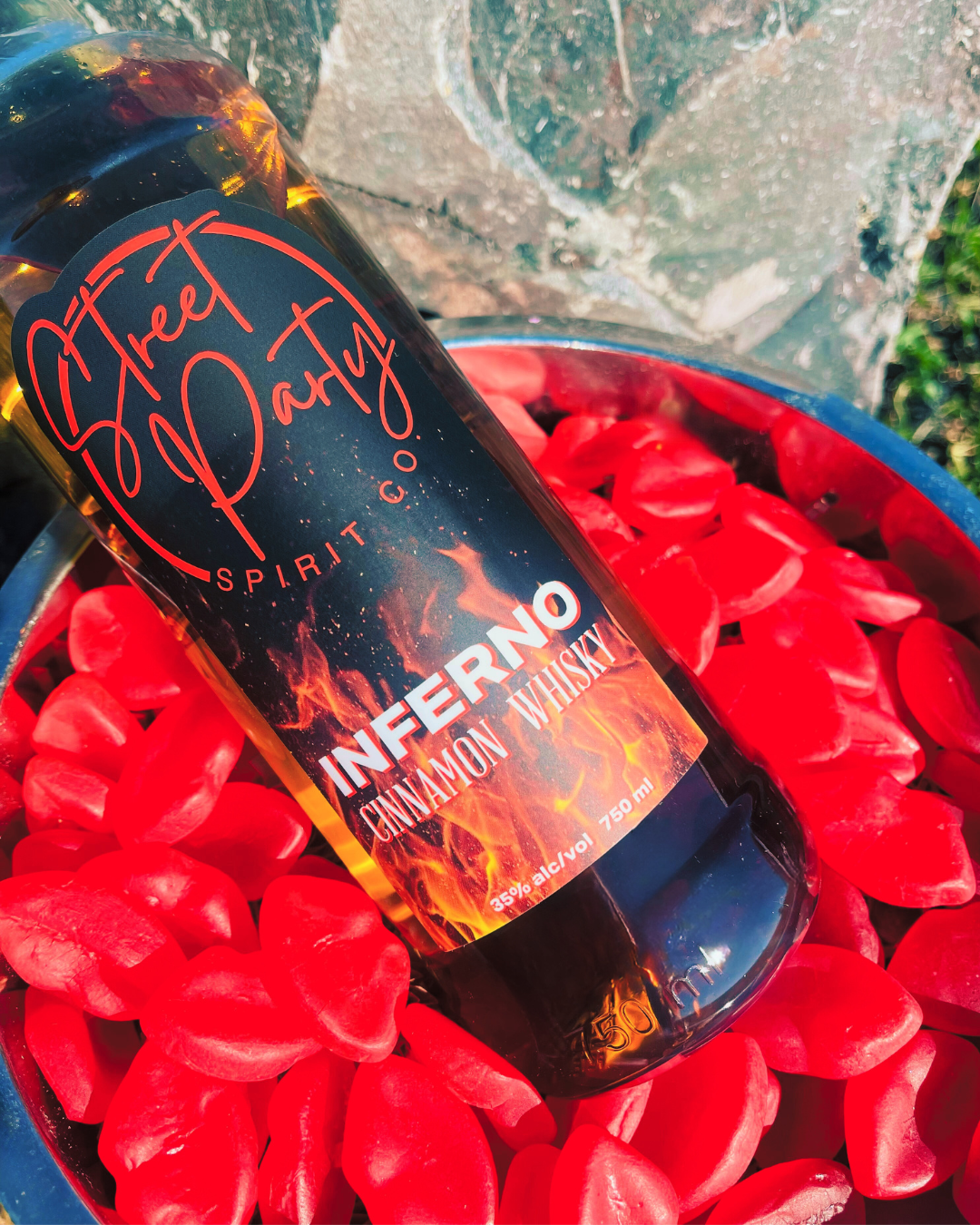 Bottle of Street Party Spirit Co. Inferno Cinnamon Whiskey surrounded by red lip candies in a metal bucket with a rock as the background