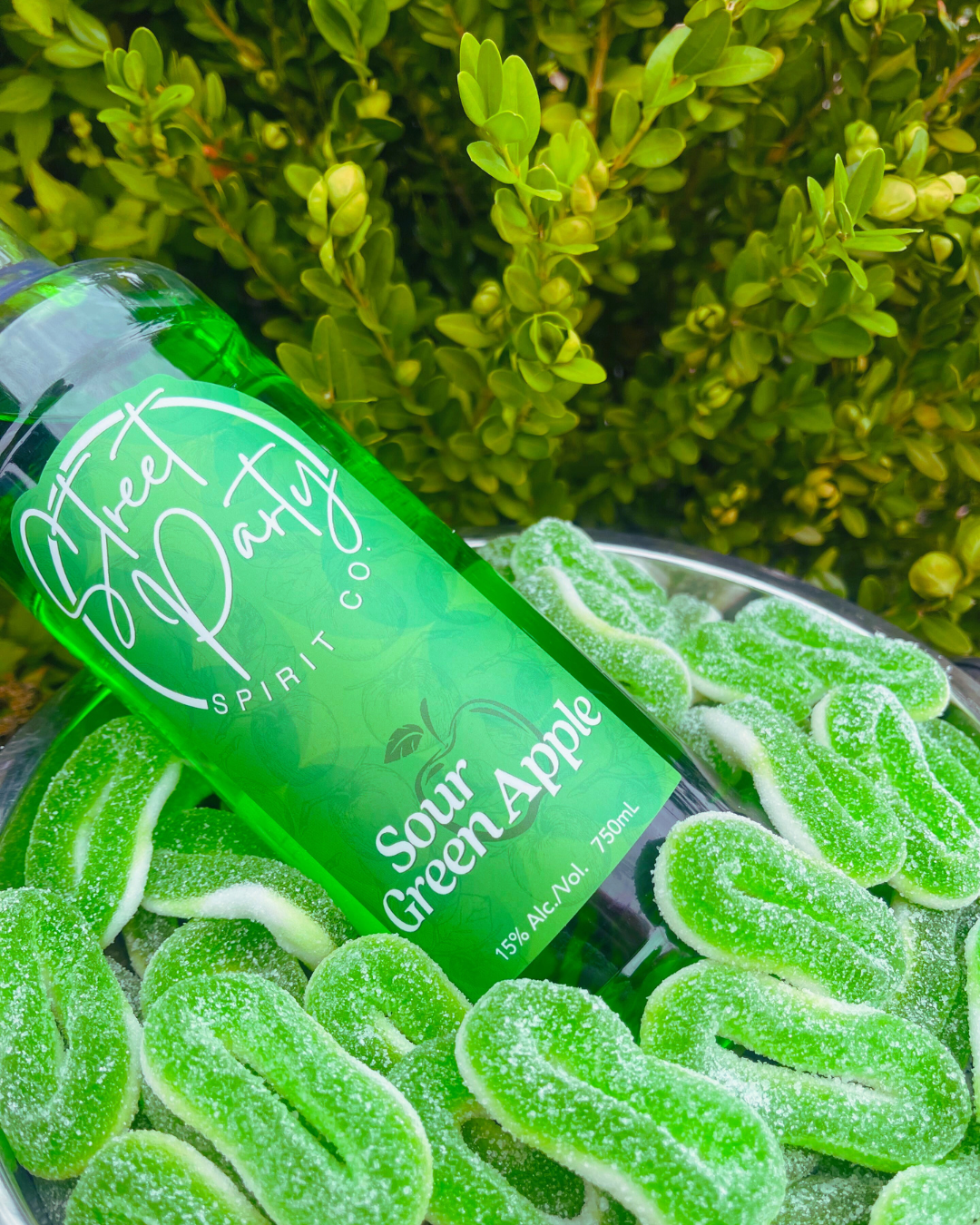 Bottle of Street Party Spirit Co. Sour Green Apple with green gummy candies in a metal bucket with a green leafy background.