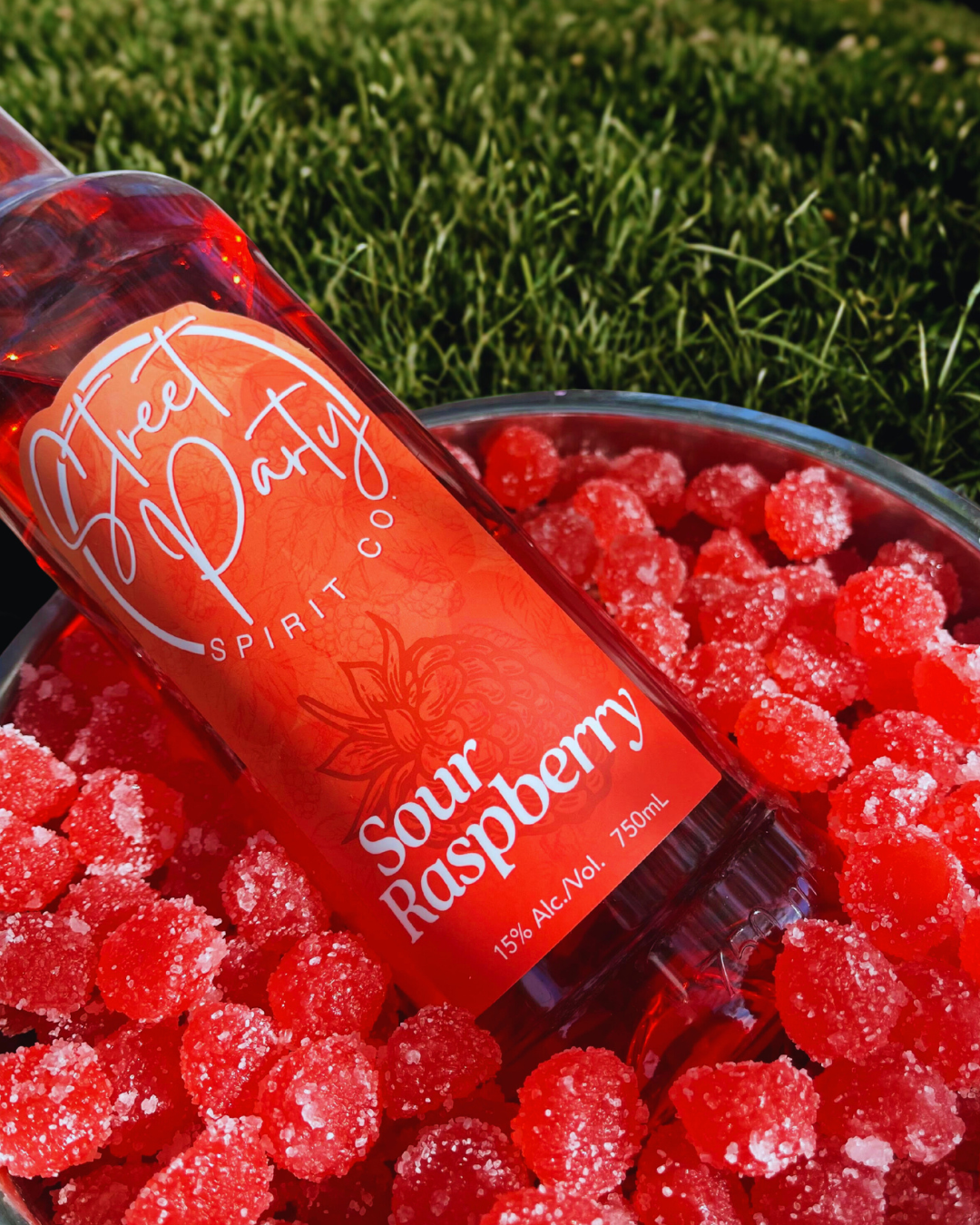 Bottle of Street Party Spirit Co. Sour Raspberry with red sour gummy candies in a metal bucket on grass.